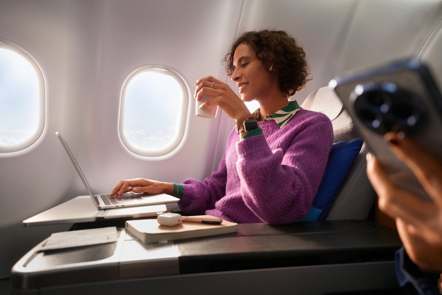 Eine Frau sitzt in der Business Class, schaut auf ihren Laptop und hat ein Getränk in der Hand