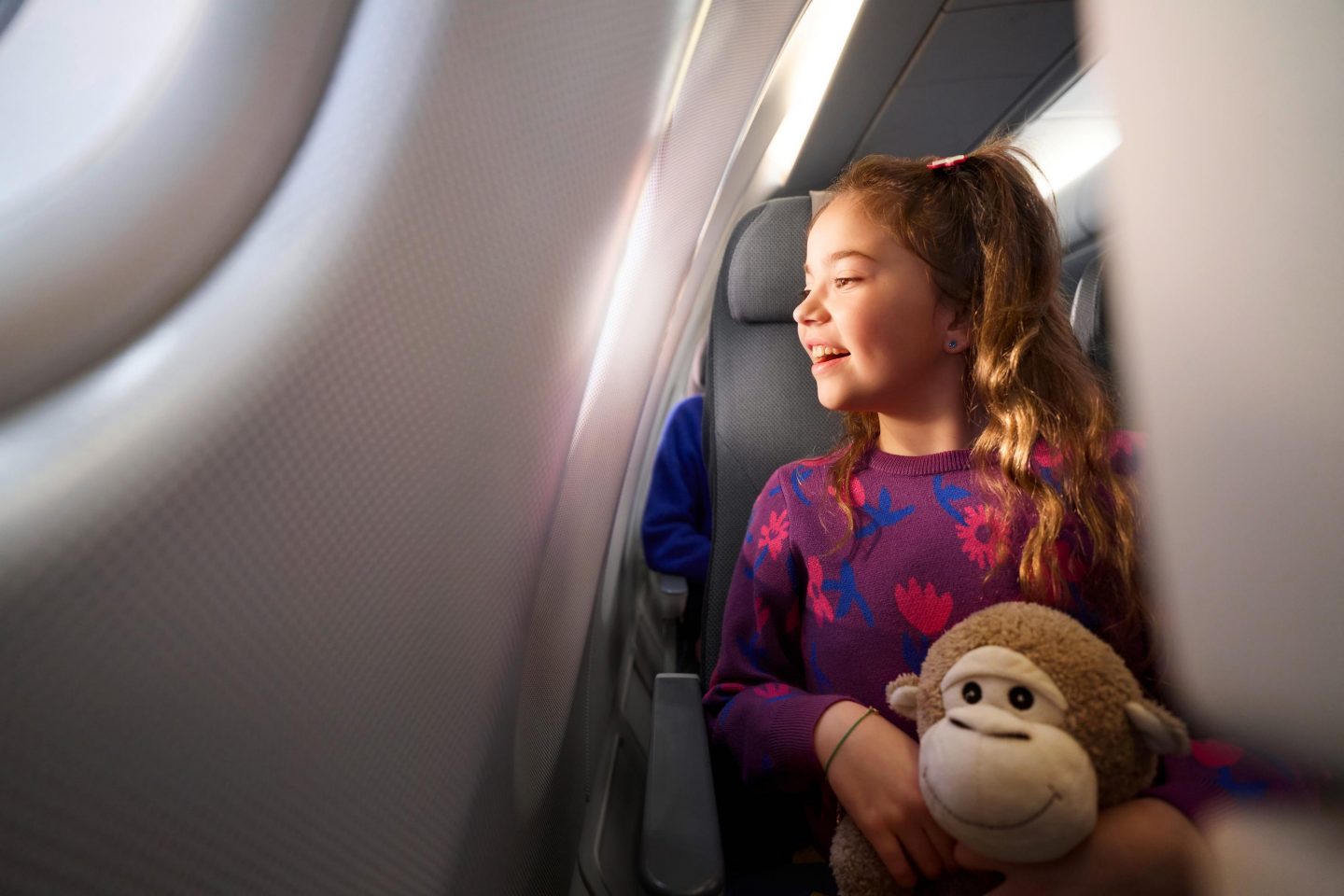 A child looks out of the airplane window with a teddy bear in her arms