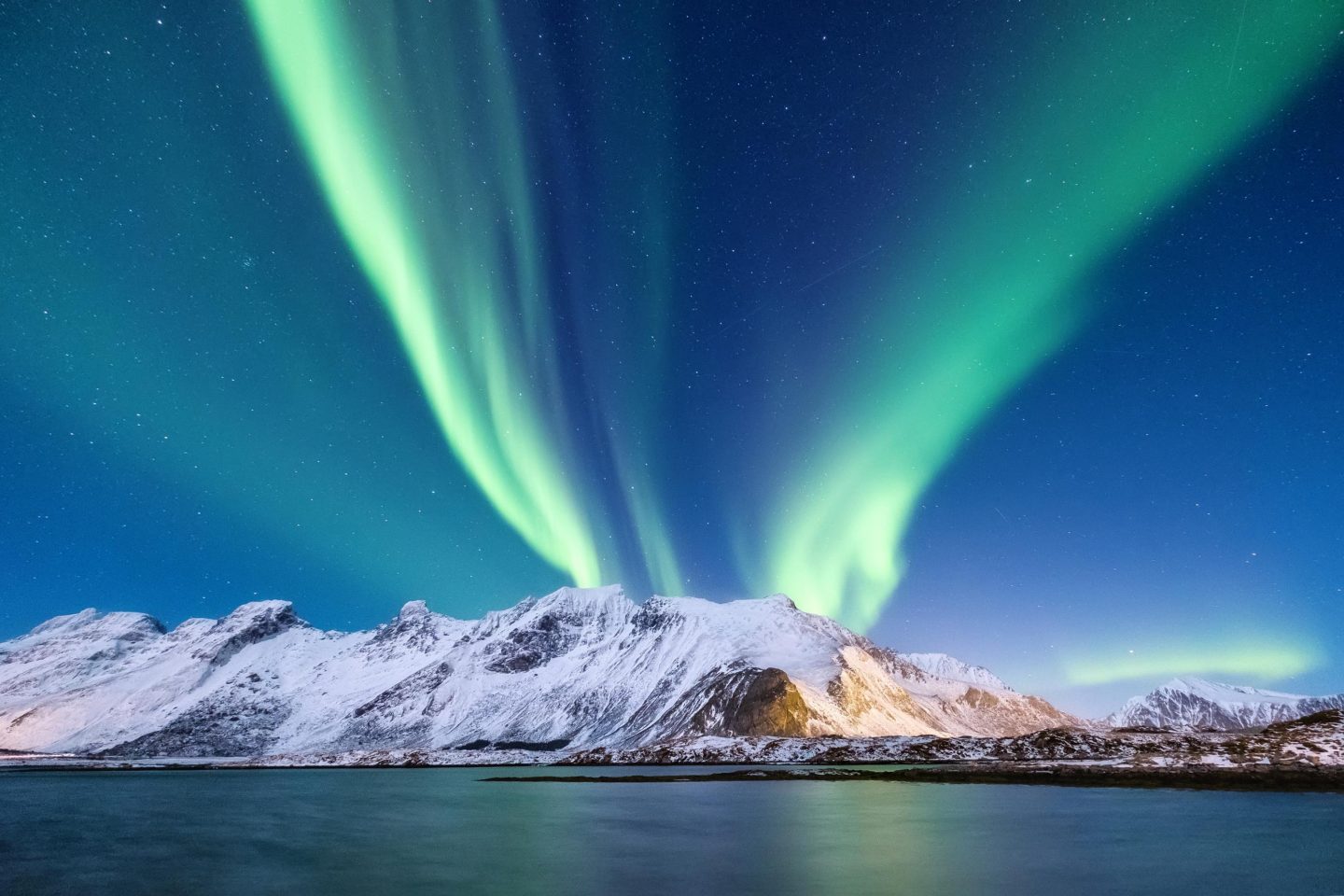 Vivid green Northern Lights illuminate the night sky above the snow-covered mountains of Evenes, Northern Norway, with calm water in the foreground and a clear starry sky overhead.