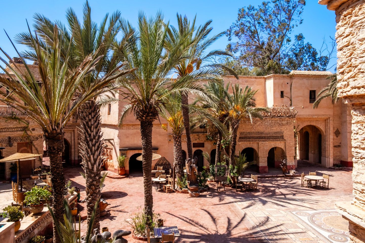 Sunny courtyard with several palm trees, light-colored Moroccan-style buildings, seating areas, and shaded archways.