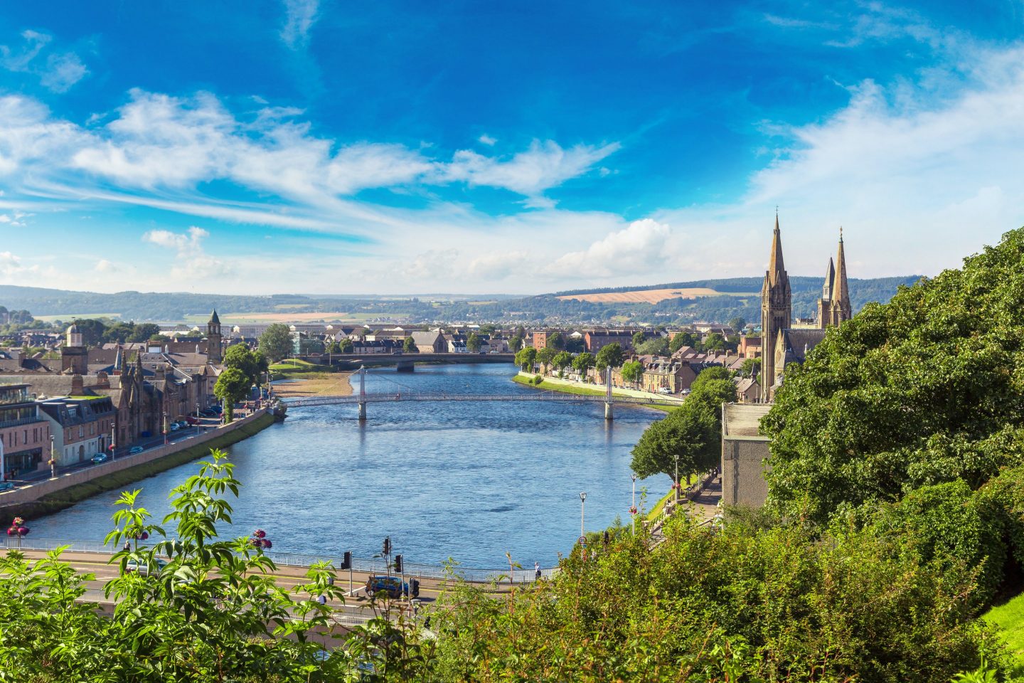 Elevated view of Inverness showing a wide river, churches with tall spires, abundant trees, and wooded hills beneath a bright blue sky.