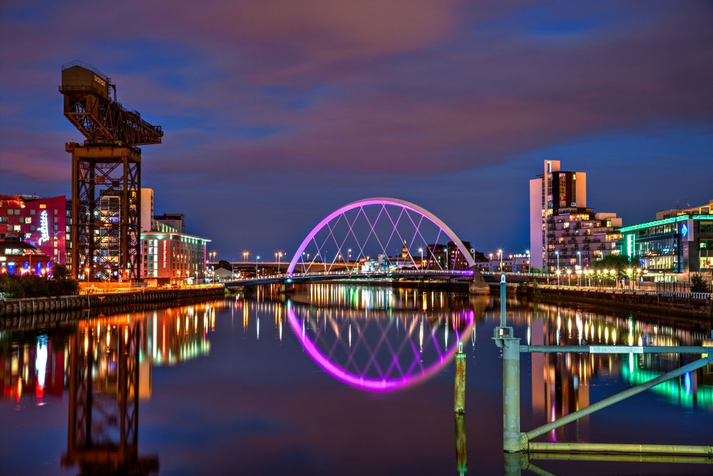 Nighttime view of the River Clyde in Glasgow with a lit arch bridge, modern riverside buildings, and reflections on the water.