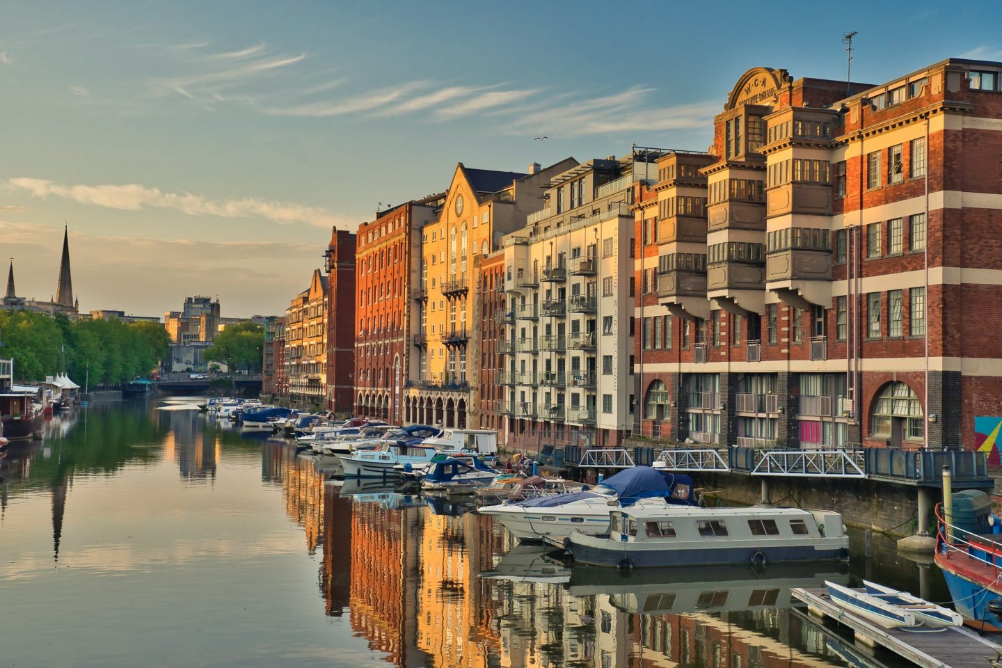 Bristol waterfront with a row of multi-story buildings, several boats moored along the quay, and reflections of the facades in the still harbor water.