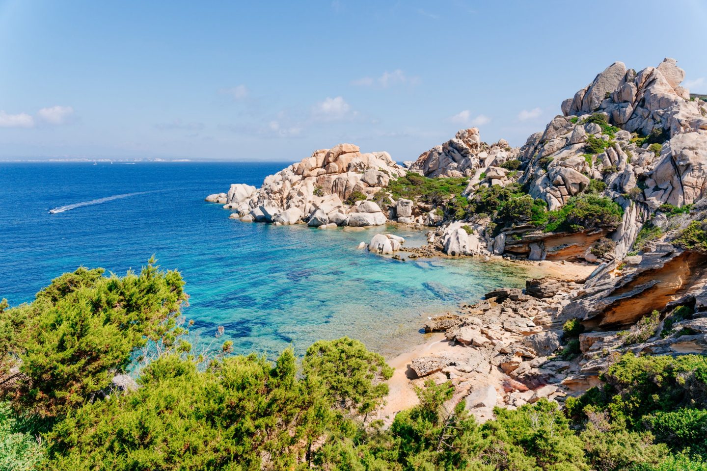 Turquoise sea in a rocky cove near Alghero, Sardinia, surrounded by Mediterranean vegetation and distinctive granite cliffs under a clear blue sky.