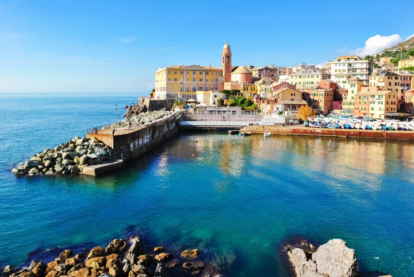 Coastal view of Genoa with colorful buildings, a small harbor, a breakwater, and clear turquoise water along the shoreline.