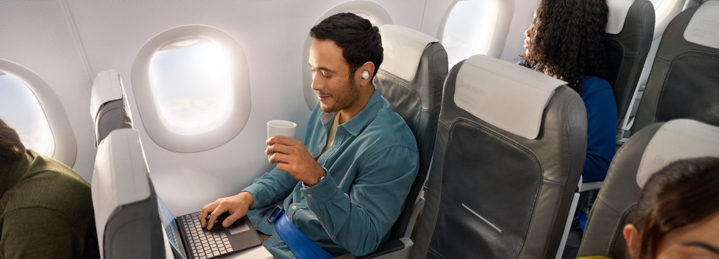 Ein Mann sitzt entspannt in der Business Class , hält einen Kaffee in der Hand und schaut auf seinen Laptop