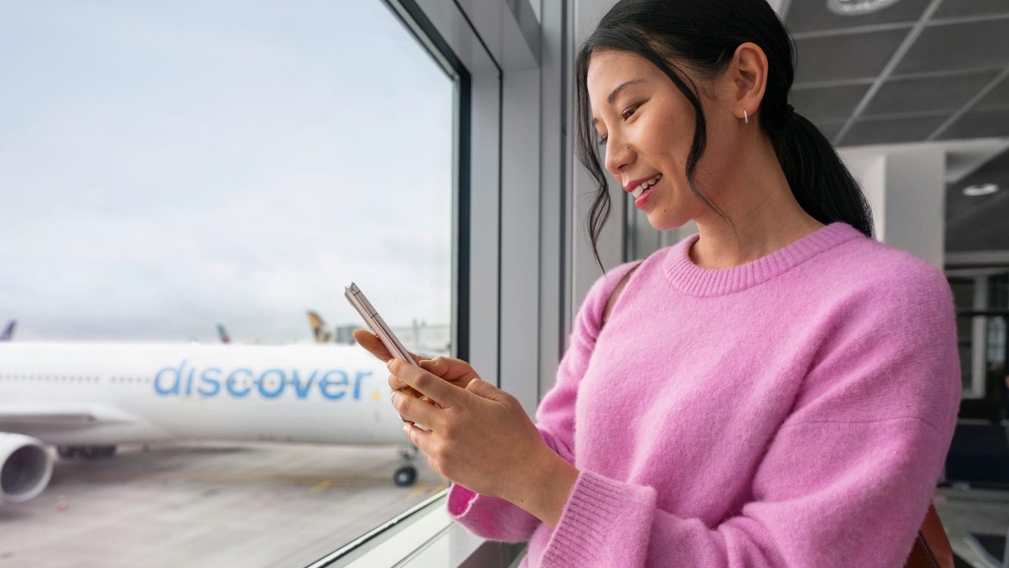 Frau mit schwarzen Haaren und pinkfarbenem Pullover am Flughafen mit einem Handy in der Hand und einem Discover Airlines Flugzeug im Hintergrund