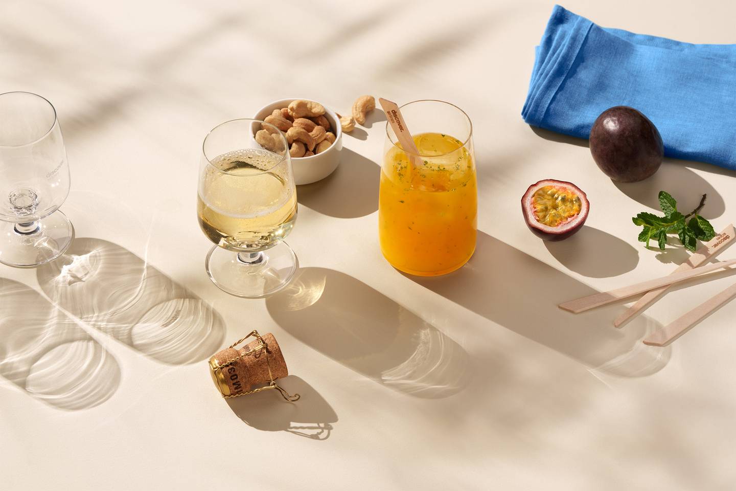 Ein Glas Sekt, ein oranger Bay Cocktail und eine kleine Schale mit Nüssen liegen auf einem Tisch